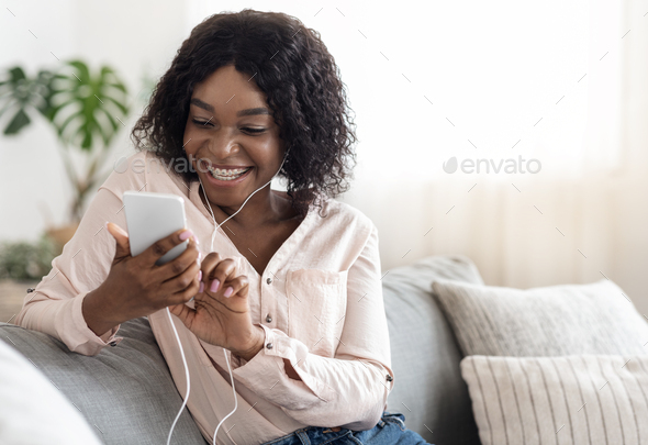 Cheerful Black Girl Messaging On Smartphone And Listening Music At Home, Relaxing On Comfortable Couch, Free Space