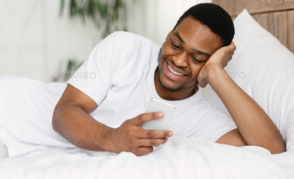 Cheerful Black Guy Using Smartphone Lying In Bed At Home On Weekend Morning. Mobile App Concept