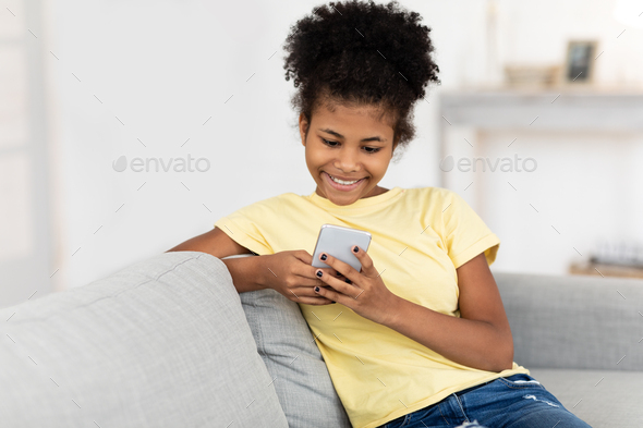 Happy Teen Girl Using Mobile Phone Chatting In Social Media Sitting On Sofa At Home. Free Space For Text
