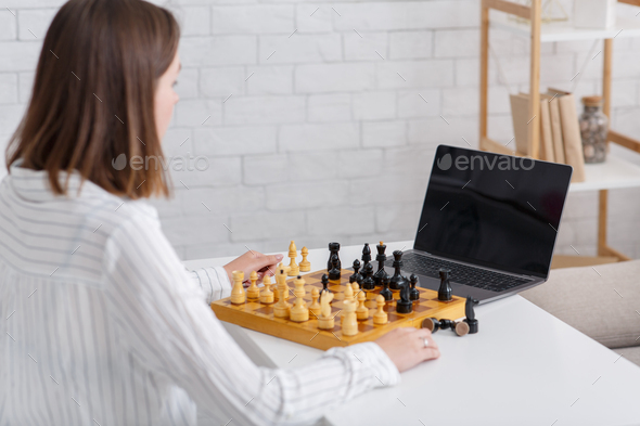 Online teacher during self-isolation at pandemic. Girl watches video lesson and play in chess in interior of living room