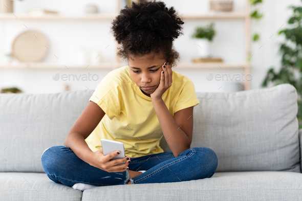 Bored African American Teen Girl Using Smartphone Browsing Internet Sitting On Sofa At Home. Modern Boring Childhood