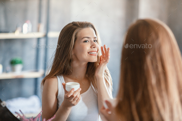Skin care routine. Pretty blonde girl applying facial cream near mirror at bathroom