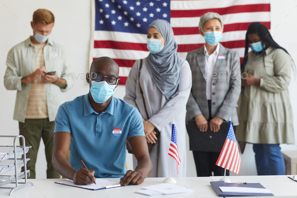 Multi-ethnic group of people standing in row and wearing masks at polling station on election day, focus on African-American man registering for voting