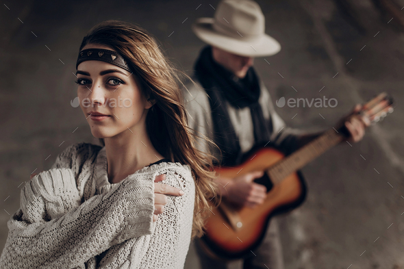 Beautiful sensual indie girl in hipster boho clothes posing in front of handsome cowboy guitar player outdoors
