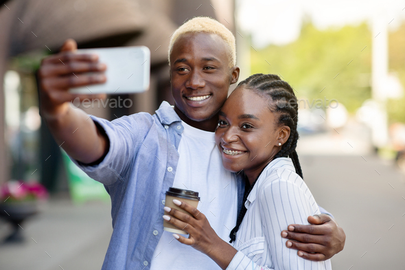 Anti-stress shopping. Happy african american millennial guy and girl with coffee, hugs and maks selfie, on street, copy space