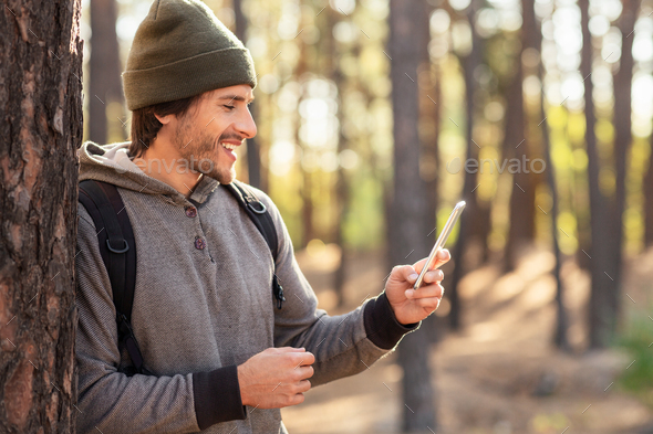 Positive guy with backpack leaning on tree in forest, using mobile phone, copy space