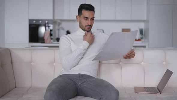 Focused Bearded Young Man Analyzing Business Graph Sitting on Couch in Home Office alt
