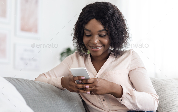 Joyful Black Woman Using Mobile Phone While Relaxing On Sofa At Home, Messaging With Friends Or Browsing Social Media, Copy Space