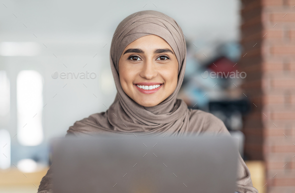 Portrait of pretty muslim girl in hijab using laptop at cafe, smiling at camera
