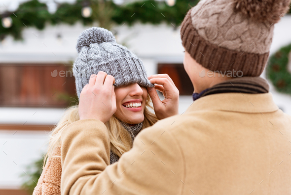 Romantic couple having fun outdoors in winter day, loving man playing with girlfriend’s hat, covering her eyes and laughing, closeup