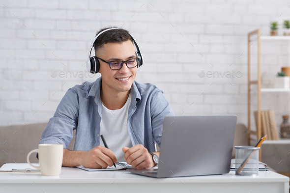 New knowledge online at home. Happy man in glasses and headphones makes notes and looks at laptop