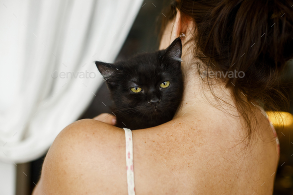 Adorable black kitten with yellow eyes on female shoulder in room. Woman hugging cute scared black cat, adoption concept. Kitty face closeup