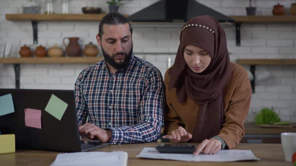 Concentrated Middle Eastern Wife Calculating Family Budget As Focused Husband Working Online on alt