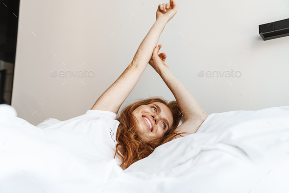 Image of ginger happy woman with freckles stretching her body while lying in bed after sleep at home