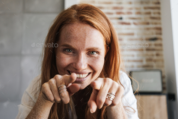 Image of happy ginger woman with freckles smiling and pointing fingers at camera in home