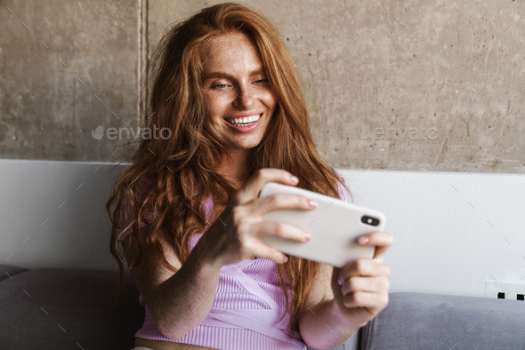 Image of redhead joyful woman playing online game on cellphone while sitting on couch at home