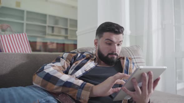 Handsome Caucasian Man Lying on Sofa and Using Tablet As His Wife Passing By with Shopping Bags alt