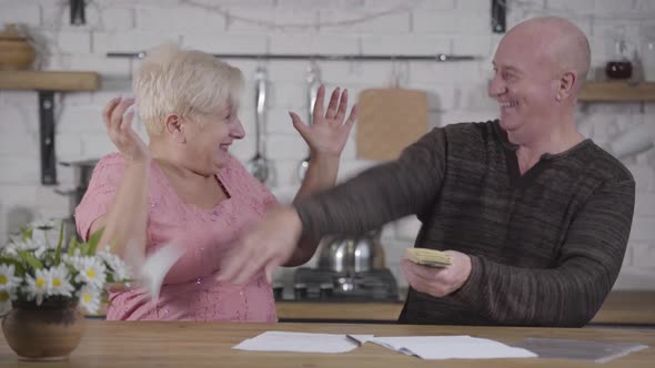 Smiling Senior Caucasian Man Showing Pack of Banknotes To His Shocked Wife. Mature Woman Throwing alt