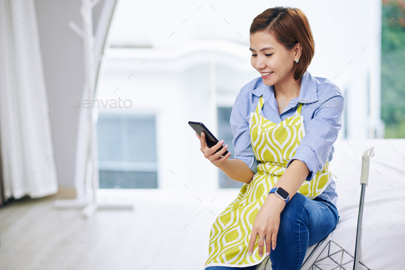 Smiling pretty Vietnamese housewife sitting on bed edge and checking text messages in her smartphone