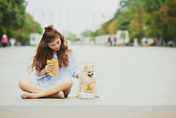 Pretty young woman and her dog sitting outdoors and having snacks