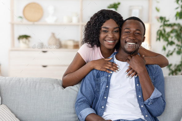 Happy african woman hugging her husband from behind, spending time together at home, empty space