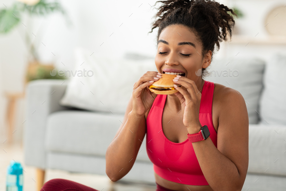 Cheat Meal. Black Fit Woman Eating Burger Enjoying Unhealthy Fast Food After Training Sitting On Floor At Home. Free Space