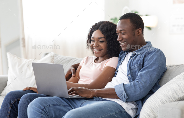 Cheerful black couple using laptop while resting on couch at living room at home, watching funny content together, copy space