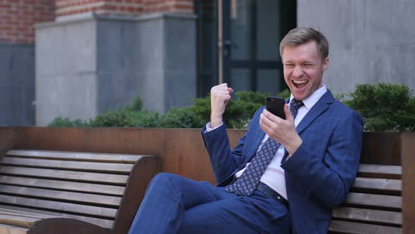 Excited Businessman Celebrating Success, while Using Smartphone alt