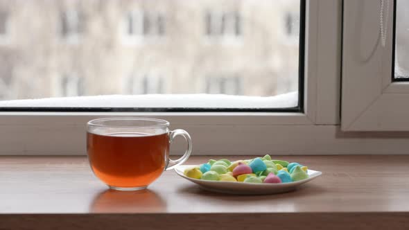 A Large Mug of Tea and Colorful Sweets in a Plate on the Windowsill By the Window in Winter