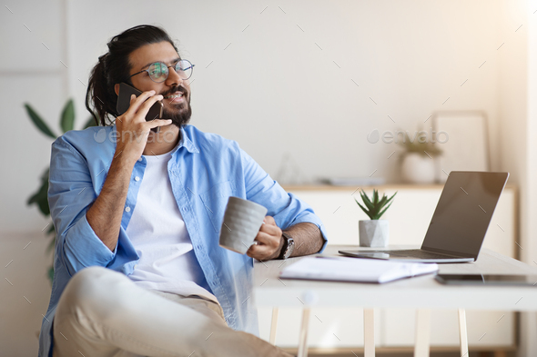 Indian Freelancer Guy Talking On Cellphone And Drinking Coffee At Home Office - Stock Photo - Images