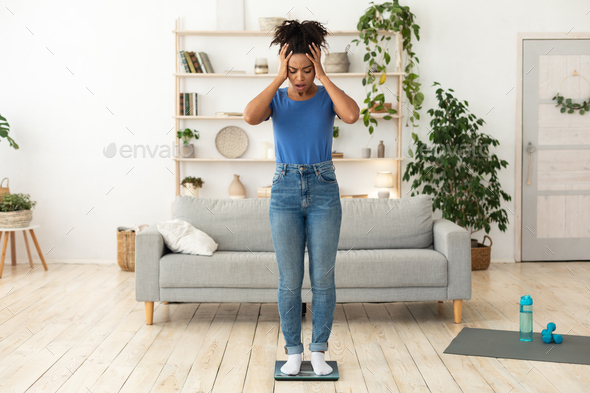 Weight Gain. Unhappy Black Girl Standing On Scales Touching Head Having Excess Kilograms Posing Indoors. Full-Length, Blank Space