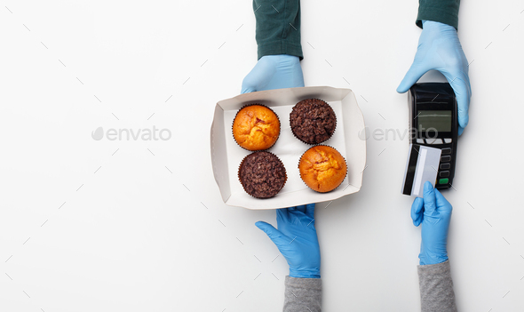 Home delivery of sweets during quarantine. Client pays for order with credit card, courier in rubber gloves gives contactless terminal and box with muffins isolated on white background, top view