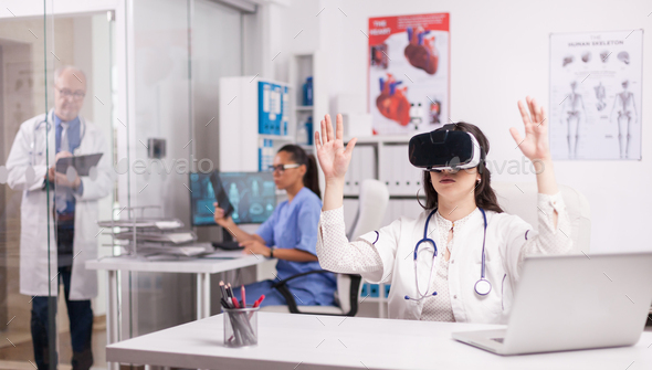 Young medic doing hand gesture wearing virtual reality headset in hospital office. Doctor in white coat with stethoscop. Senior physician writing notes on clipboard. Nurse in blue uniform looking at x-ray.