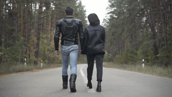 Camera Follows Happy Relaxed Millennial Couple Walking Along Empty Spring or Autumn Road. Back View alt