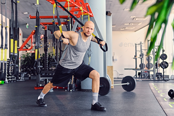 The full body image of athletic bearded tattooed, shaved head male doing trx straps exercises in a gym club.