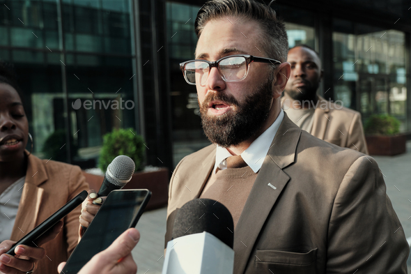 Mature bearded man in eyeglasses standing on the street among journalist and giving an interview