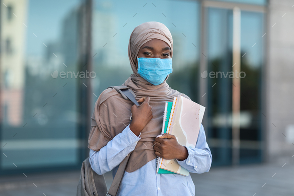 Black muslim female student wearing medical mask posing outdoors with workbooks