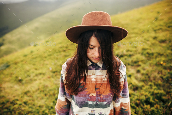 Traveler hipster girl in hat, with backpack and windy hair, smiling in mountains in clouds - Stock Photo - Images