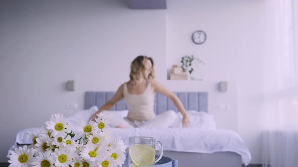 Sleepy Young Woman Wakes Up on Bed and Sits Stretching Arms