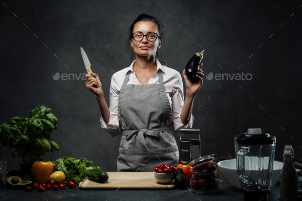 Beautiful mature woman cooking in kitchen, posing holding a knife and eggplant - Stock Photo - Images