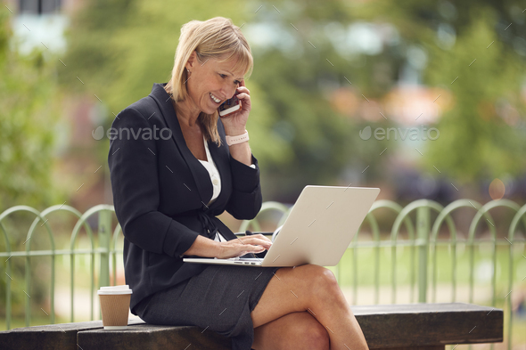 Mature Businesswoman Sitting On City Park Bench Working On Laptop And Talking On Mobile Phone - Stock Photo - Images