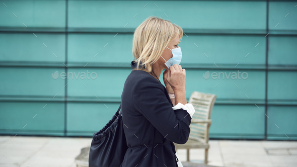 Mature Businesswoman Putting On PPE Face Mask Walking Outdoors In Street During Health Pandemic - Stock Photo - Images