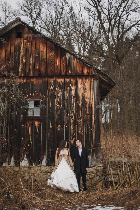 Gorgeous bride in coat and stylish groom posing at wooden house - Stock Photo - Images