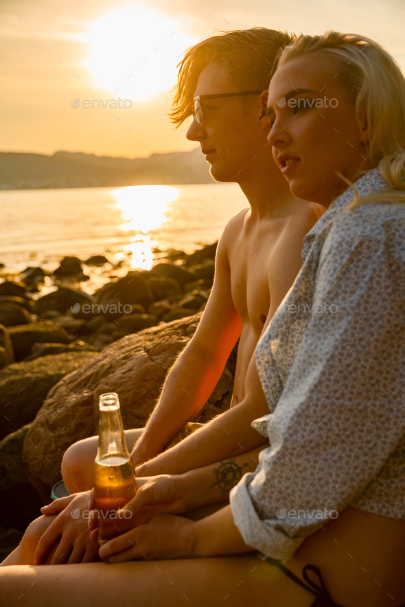 Beautiful couple enjoys their vacation while drinking beer at the beach in sunset.
