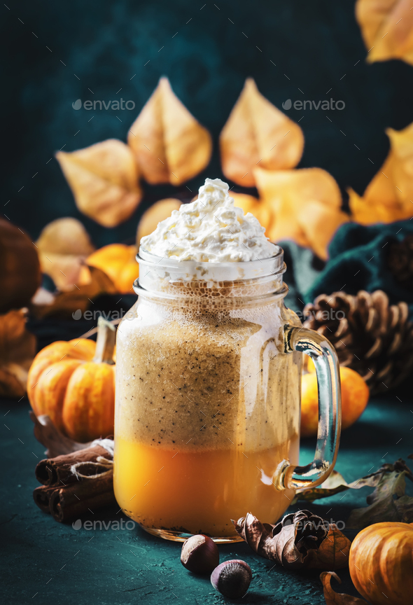 Pumpkin spiced latte or coffee in glass jar on blue table. Autumn or winter hot drink in festive natural table setting with orange leaves, spices, small pumpkins, pine cones