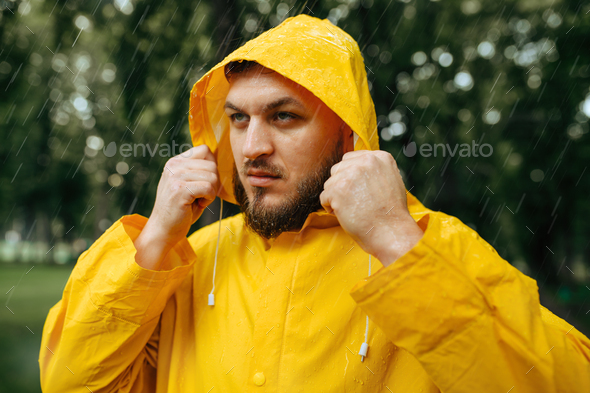 Man in raincoat puts on a hood, summer park in rainy day. Alone male person in rain cape on walking path, wet weather in alley