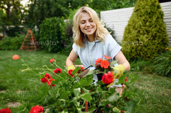 Happy young woman in gloves works with flower in the garden. Female gardener takes care of plants outdoor, gardening hobby, florist lifestyle and leisure