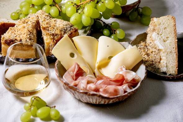 Appetizers antipasti with white sicilian focaccia. Traditional bread sliced cake with onion served with prosciutto ham, cheese, grapes and glass of white wine on white table cloth.