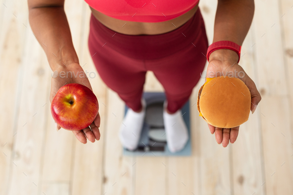 Healthy Diet And Weight Loss. Slim Black Lady Choosing Between Burger And Apple Standing On Weight-Scales At Home. High-Angle, Cropped