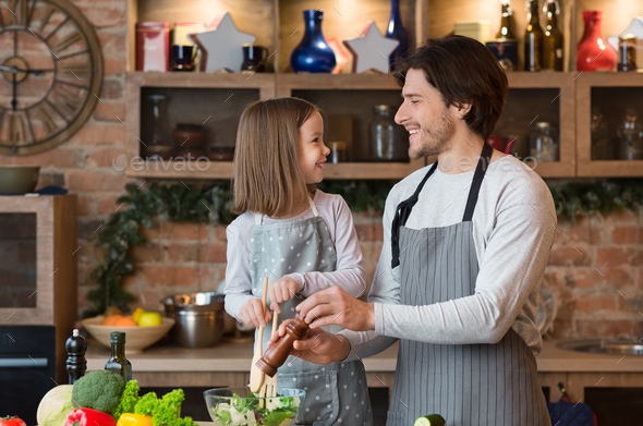 Joyful Young Dad And Little Daughter Cooking Vegetable Salad For Lunch Together, Adding Seasoning To Bowl, Enjoying Healthy Homemade Food, Having Fun At Home, Wearing Aprons And Smiling, Free Space
